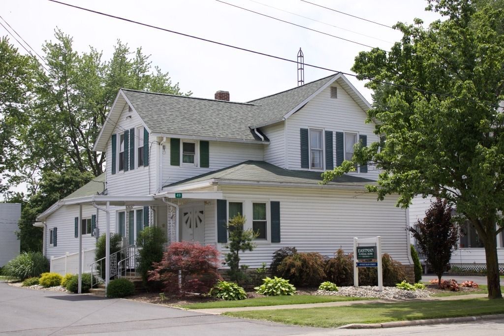 White two-story house with green shutters and landscaping, sign in front, trees in the background, daytime.