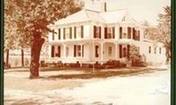 White Victorian house with black shutters, tall tree, and a grassy yard.