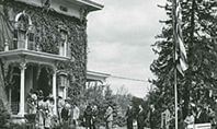 Group of people outside a large house covered in ivy, with an American flag on a pole.