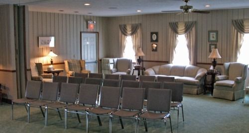A room set up for a gathering, chairs in rows face couches and windows; walls are wood paneling.