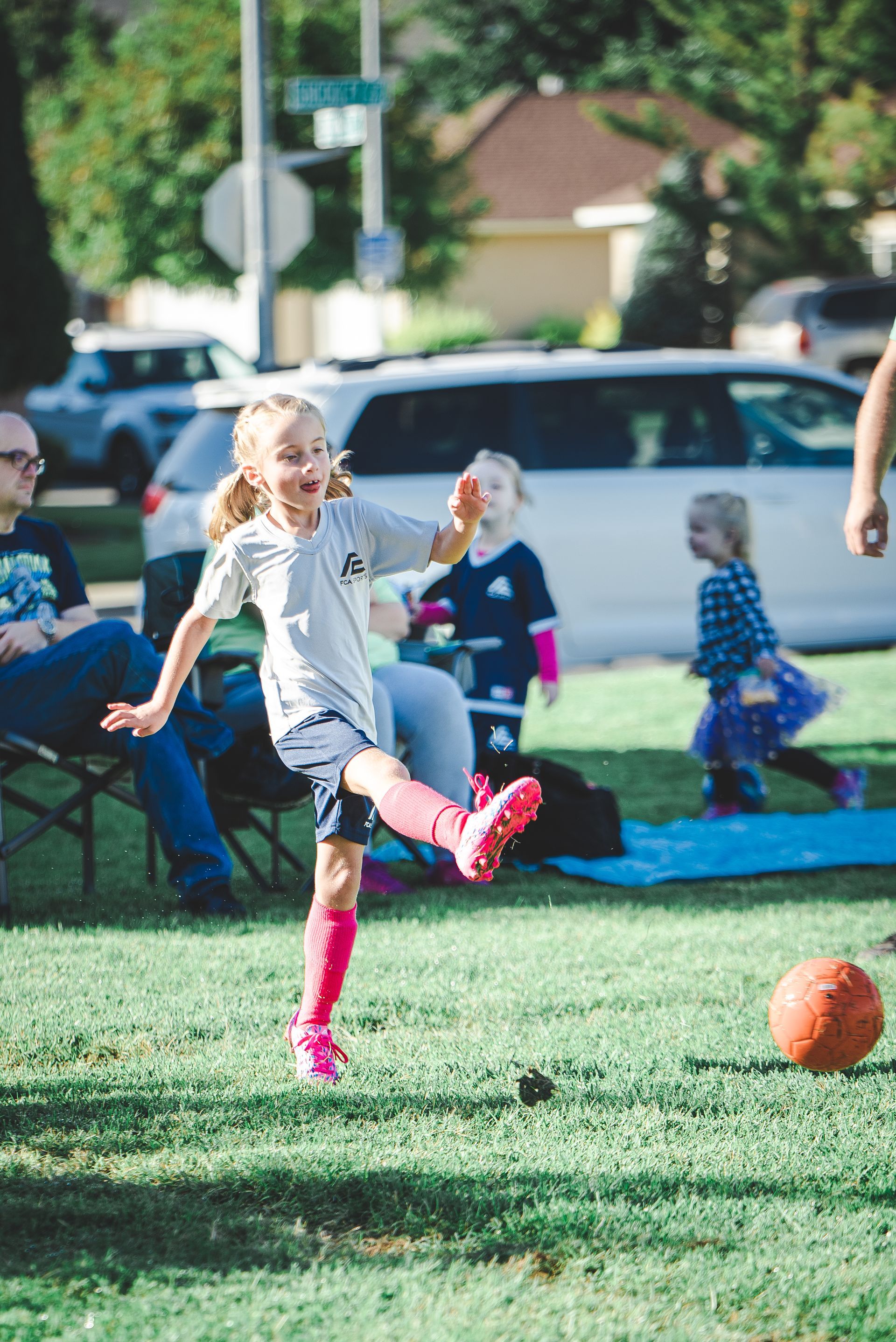 Child in a gray hoodie kicks an orange ball on a grassy field at a park