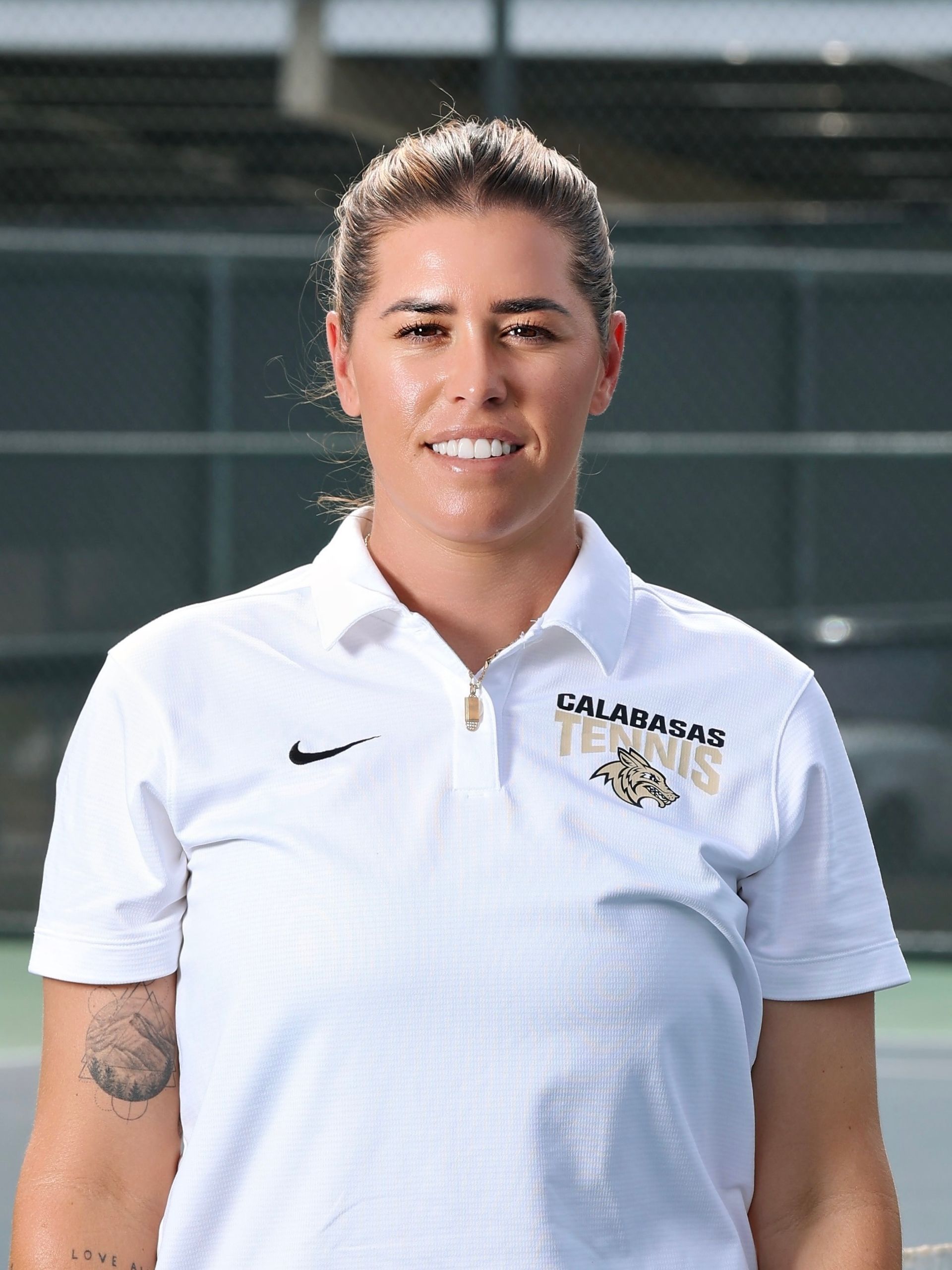 Woman with light brown hair smiling in a grey polo shirt in front of a green fence.