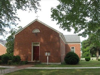A small, red brick church with a dark red door, a cross on the roof, and a semi-circular window, set among trees and lawn.