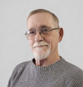 A person wearing glasses and a gray textured sweater stands against a plain, light background.