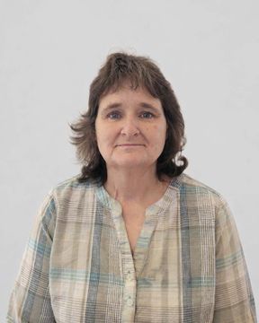 A person with shoulder-length brown hair, wearing a beige plaid shirt, poses against a plain light background.
