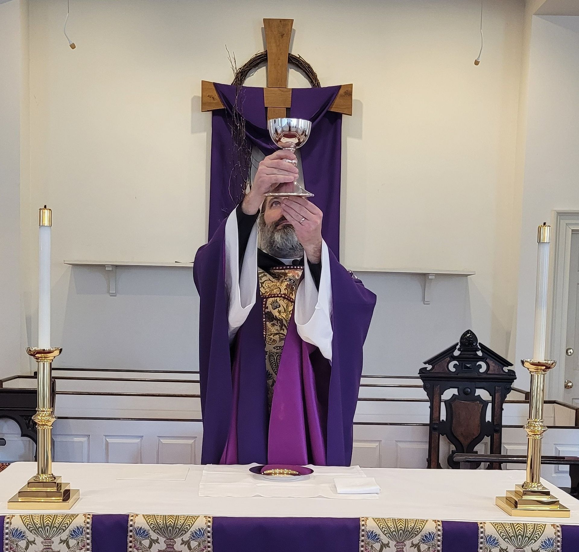 A priest in purple vestments elevates a silver chalice in front of a wooden cross draped in purple cloth on an altar.