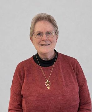 A person with short hair and glasses wearing a red sweater and gold necklace smiles against a plain white background.