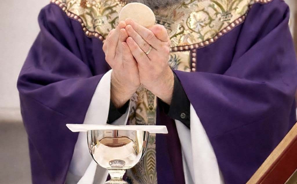 A priest in purple vestments elevates a communion host above a chalice during a Catholic Mass.