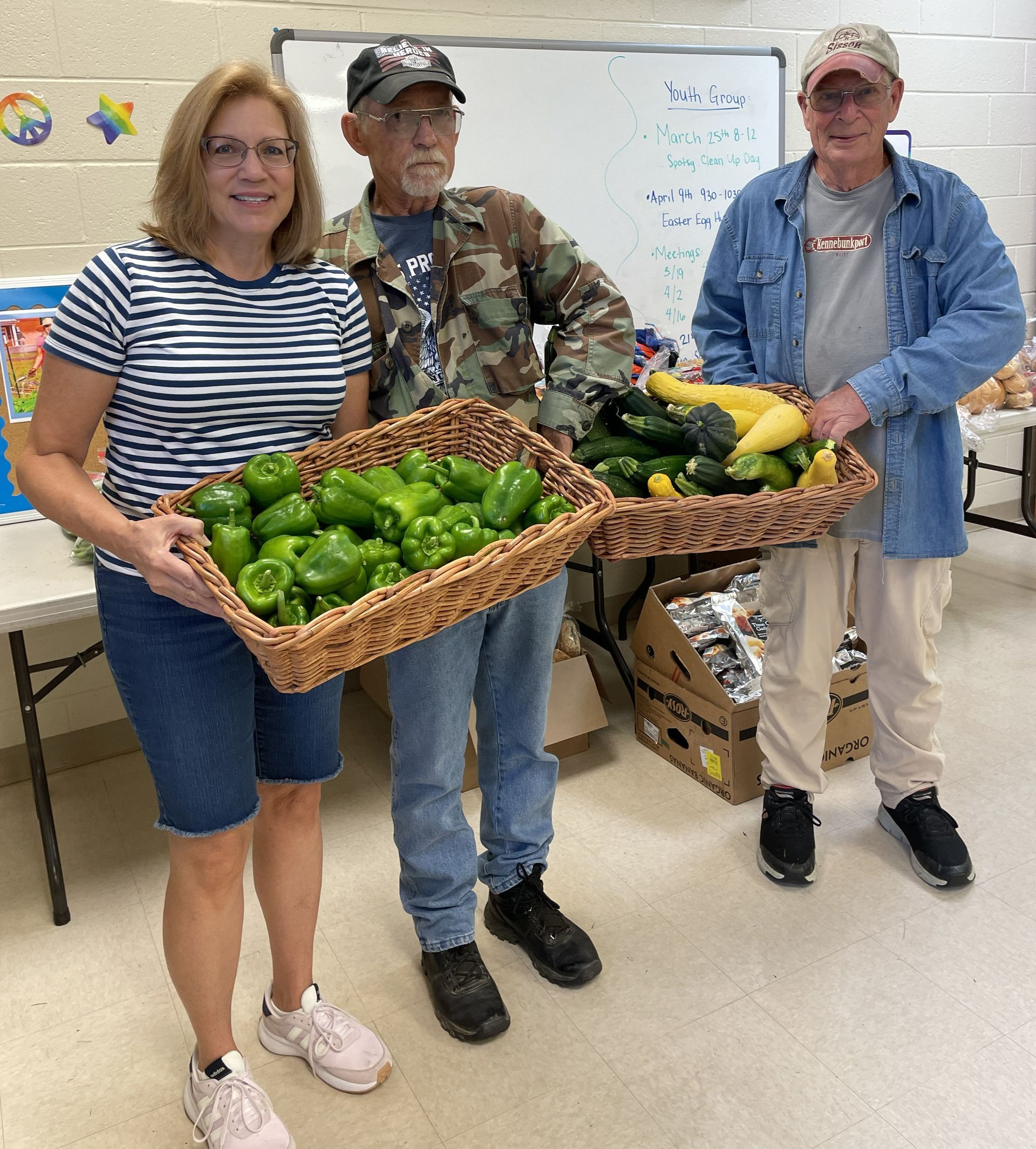 Three people stand in a room holding two wicker baskets filled with fresh green peppers and squash.