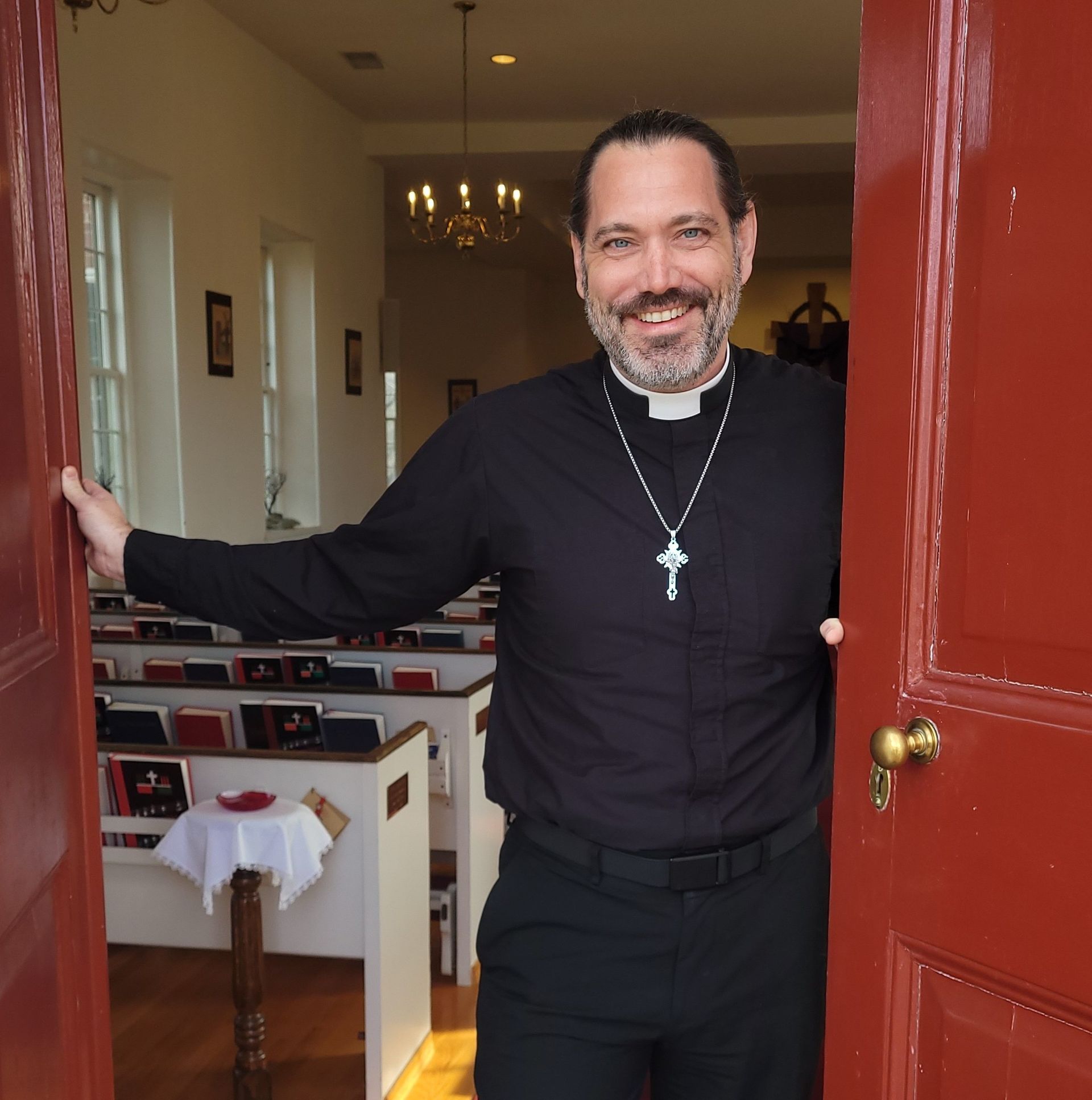 Rev Daniel in clerical clothing stands in a doorway, welcoming others into a church with pews and a chandelier.