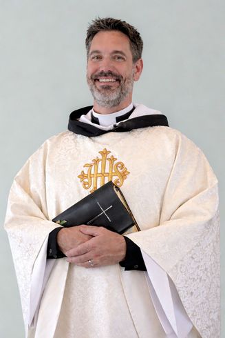 A smiling person in white liturgical vestments holds a small, black book with a cross against a plain background.