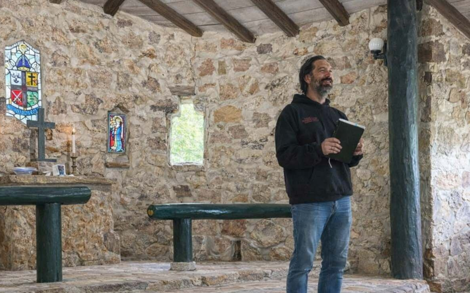 A person in a black hoodie and blue jeans holds a book while speaking inside a rustic chapel with stone walls.