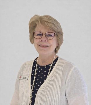 A person with blonde hair and glasses wearing a black polka-dot shirt, white cardigan, and pearl necklace.