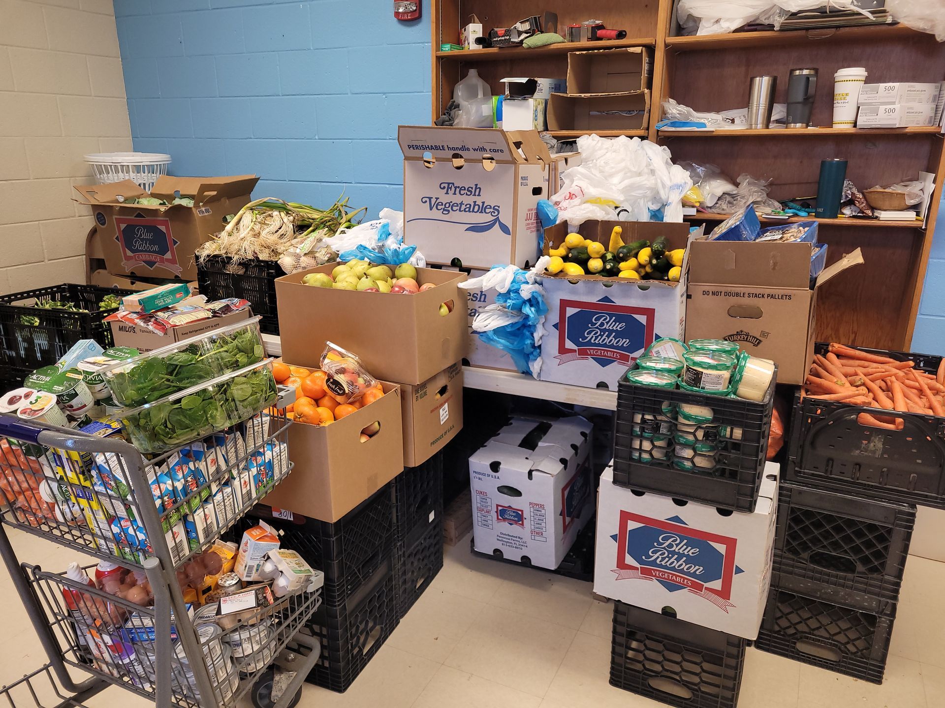 Food pantry shelves and floor packed with crates and boxes of fresh produce, including oranges, lemons, and carrots.