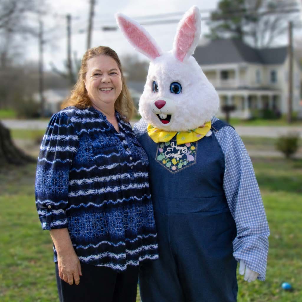 A smiling person stands next to a costumed Easter Bunny in overalls and a yellow bowtie in an outdoor setting.