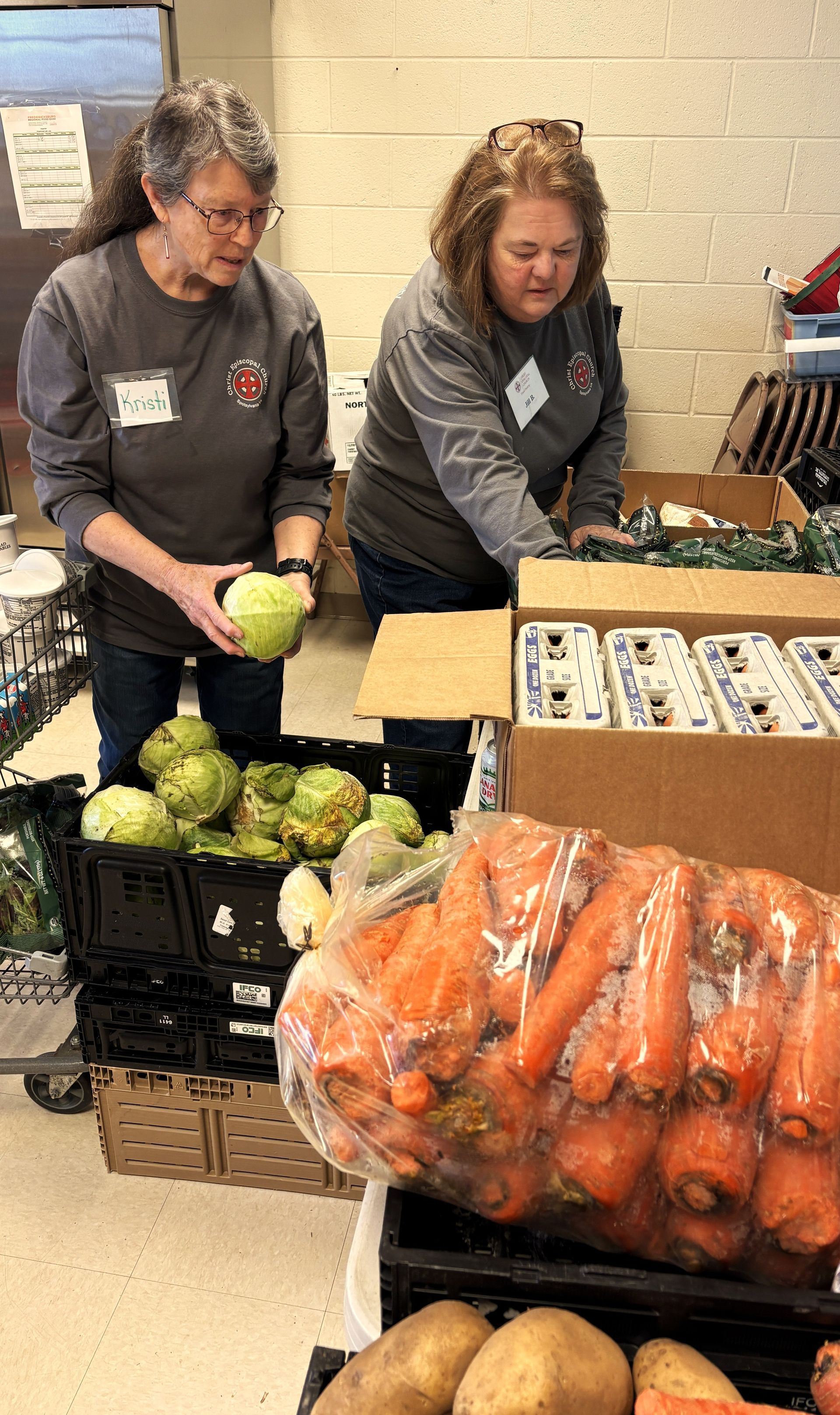 Two people in gray shirts pack fresh produce and cartons of eggs into boxes at a food distribution center.