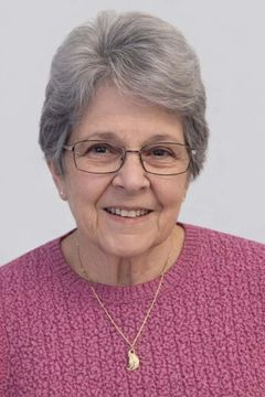 A smiling person with grey hair and glasses, wearing a pink sweater and a gold necklace, against a light grey background.