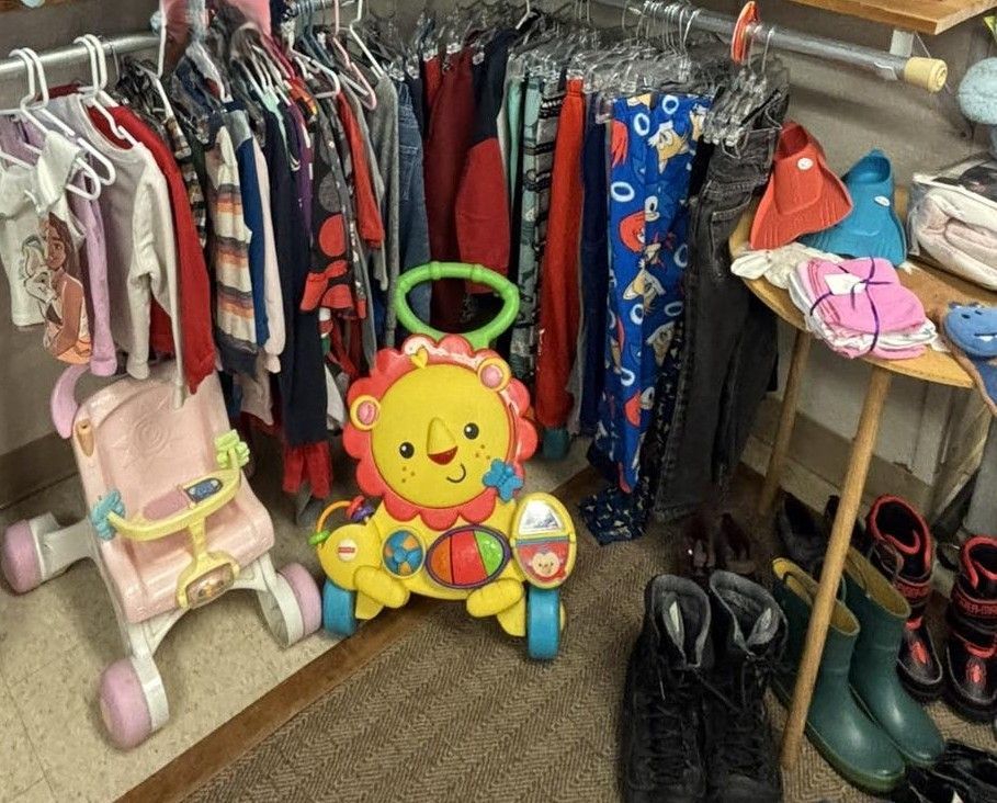 A row of children's clothes on hangers, with a lion push-walker, a small pink toy stroller, and shoes on a wooden table.