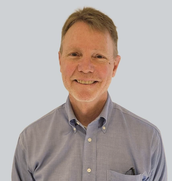 A person with short hair smiles warmly, wearing a light blue, button-down collared shirt against a gray background.