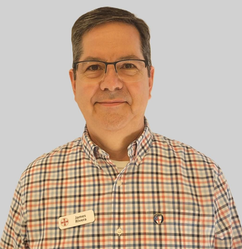 A person wearing glasses and a plaid, button-down shirt stands against a neutral background.