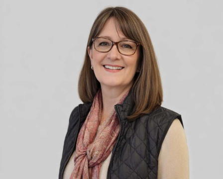 A smiling person wearing glasses, a beige sweater, a quilted black vest, and a patterned pink scarf against a gray wall.