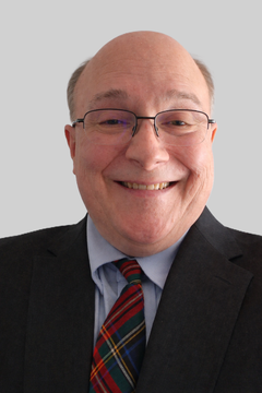 A smiling person wearing glasses, a blue dress shirt, a plaid tie, and a dark blazer against a plain gray background.
