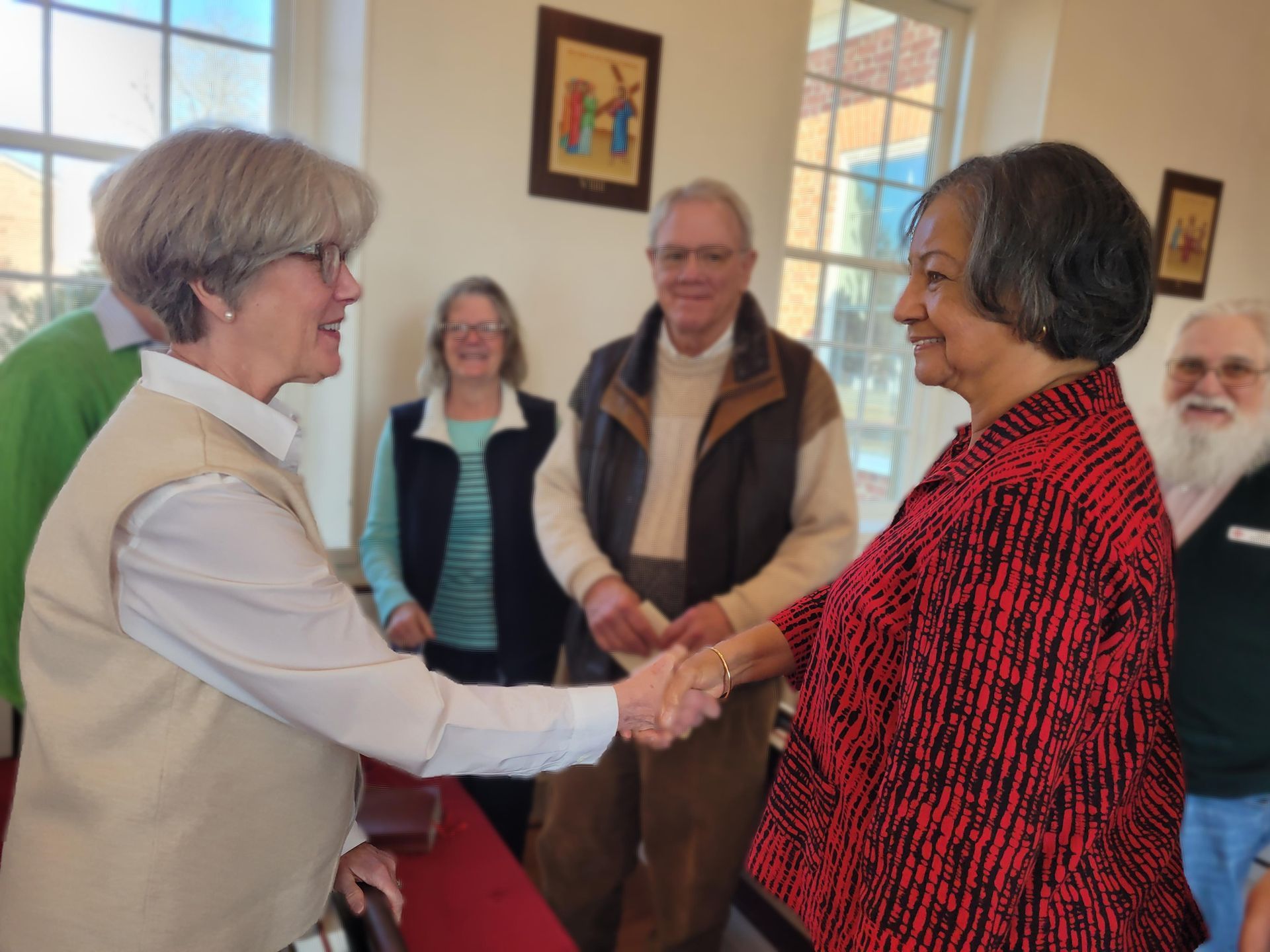 Two people shake hands in a room with others standing in the background.
