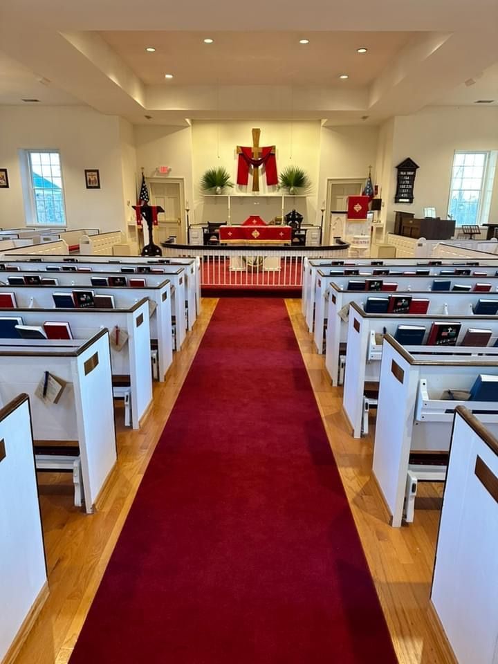 Interior of the church with white pews lining a center aisle covered by a long red carpet leading to an altar with a cross.