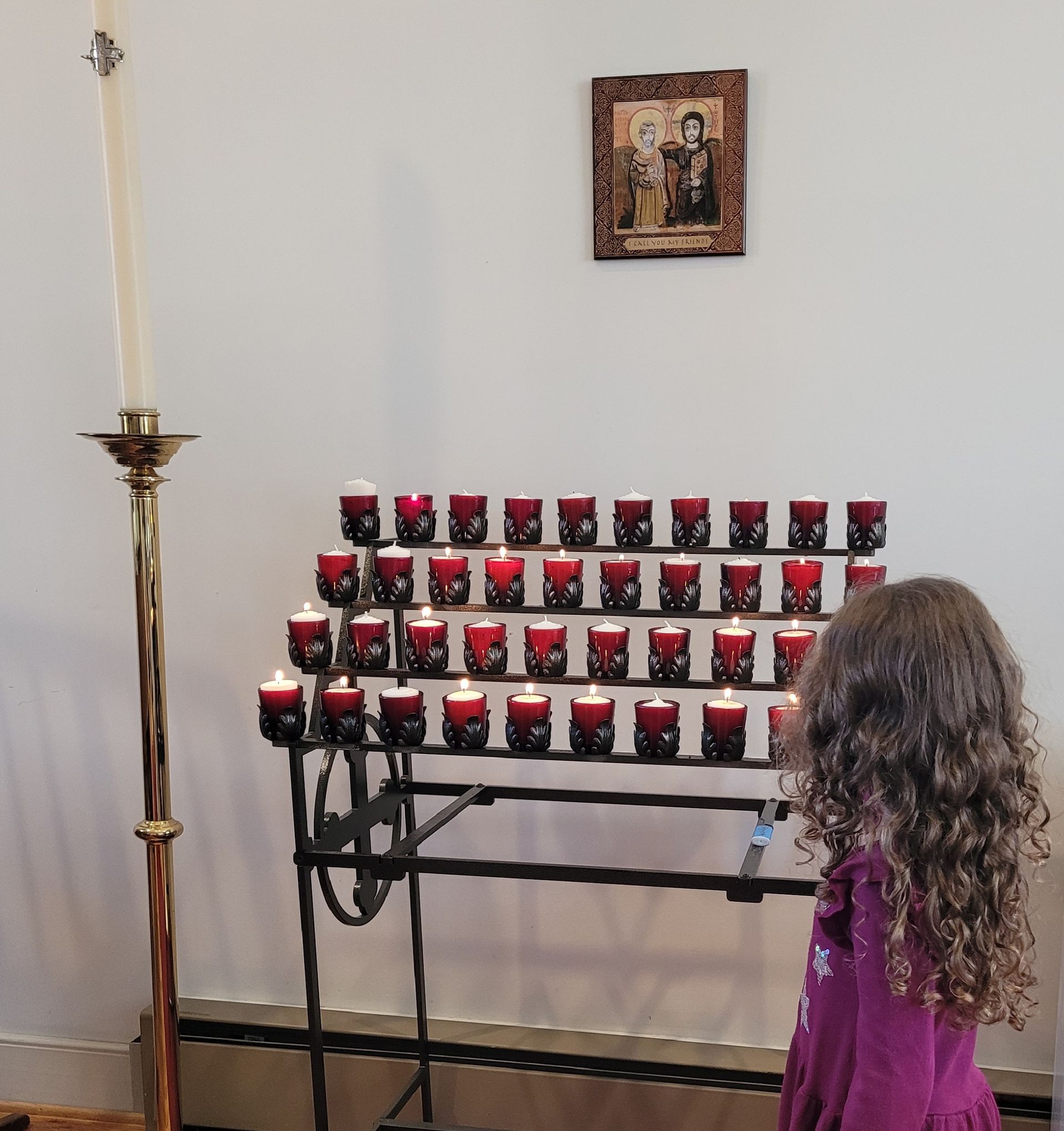 A person with long curly hair stands before a rack of lit red votive candles in a church, facing a religious icon.