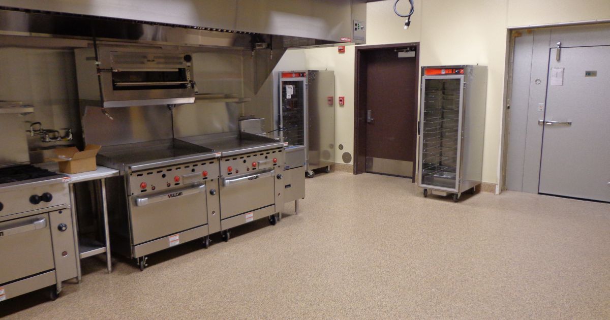Commercial kitchen with stainless steel appliances: oven, stove, and warming cabinets. A door is to the right.