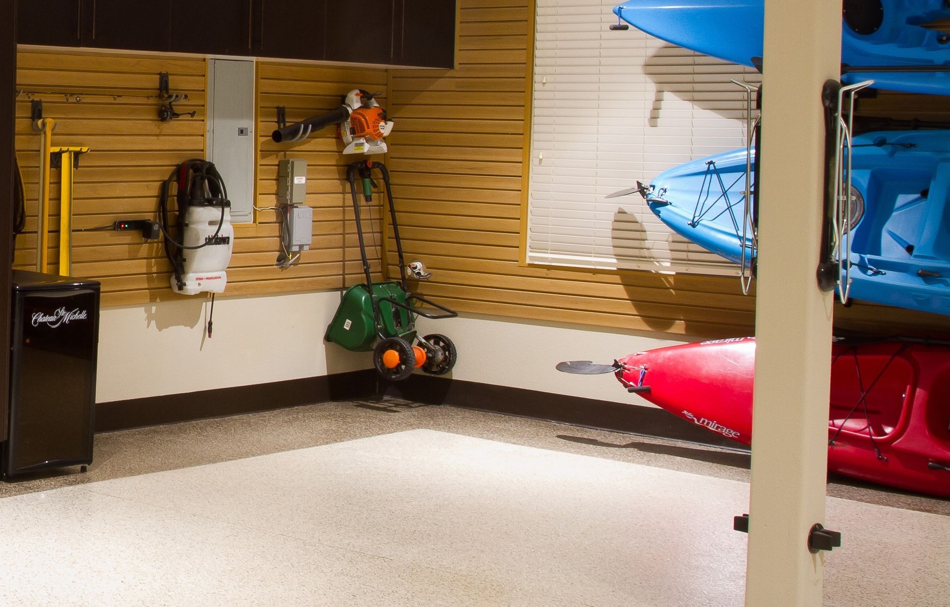 Garage with wall-mounted storage and kayaks. Tools and equipment hung on wood-slat walls. Epoxy flooring.