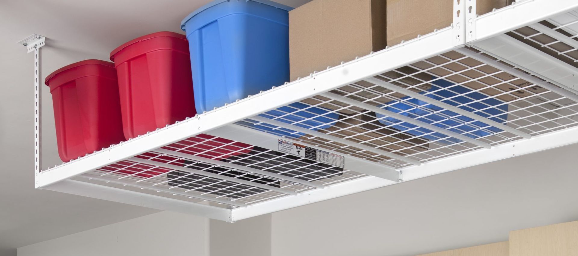 White wire-frame overhead storage shelves hold red, blue, and brown storage containers in a garage.