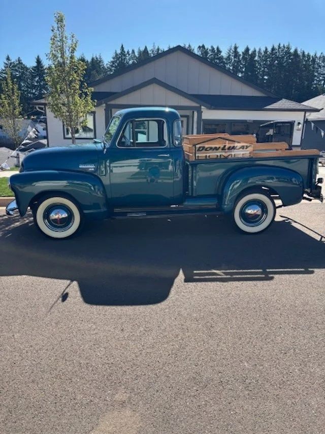 Teal vintage pickup truck parked on asphalt street with a wooden bed, white wall tires, and blue sky.