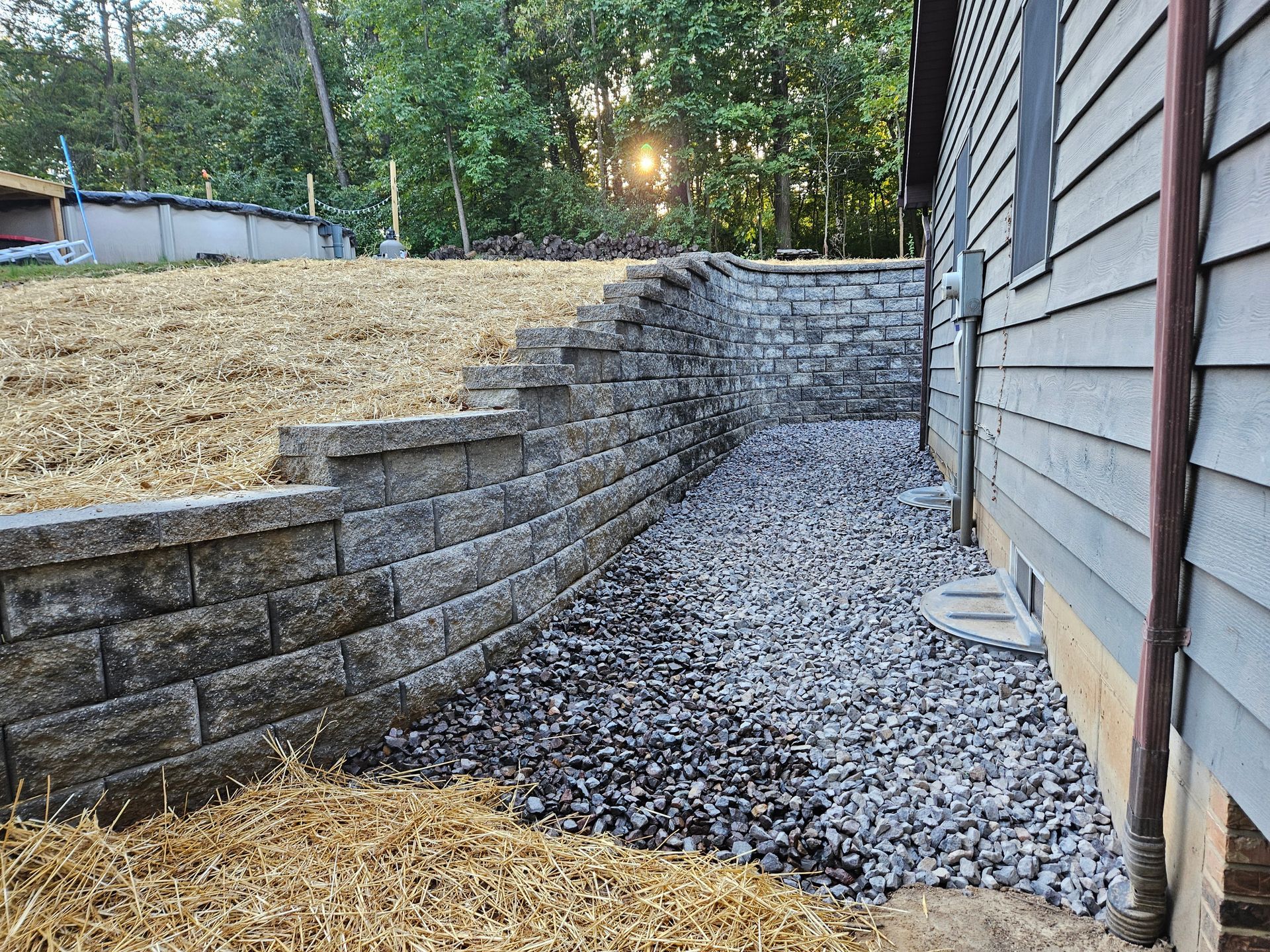 Stone retaining wall next to a house with gravel pathway, sun shining through trees.