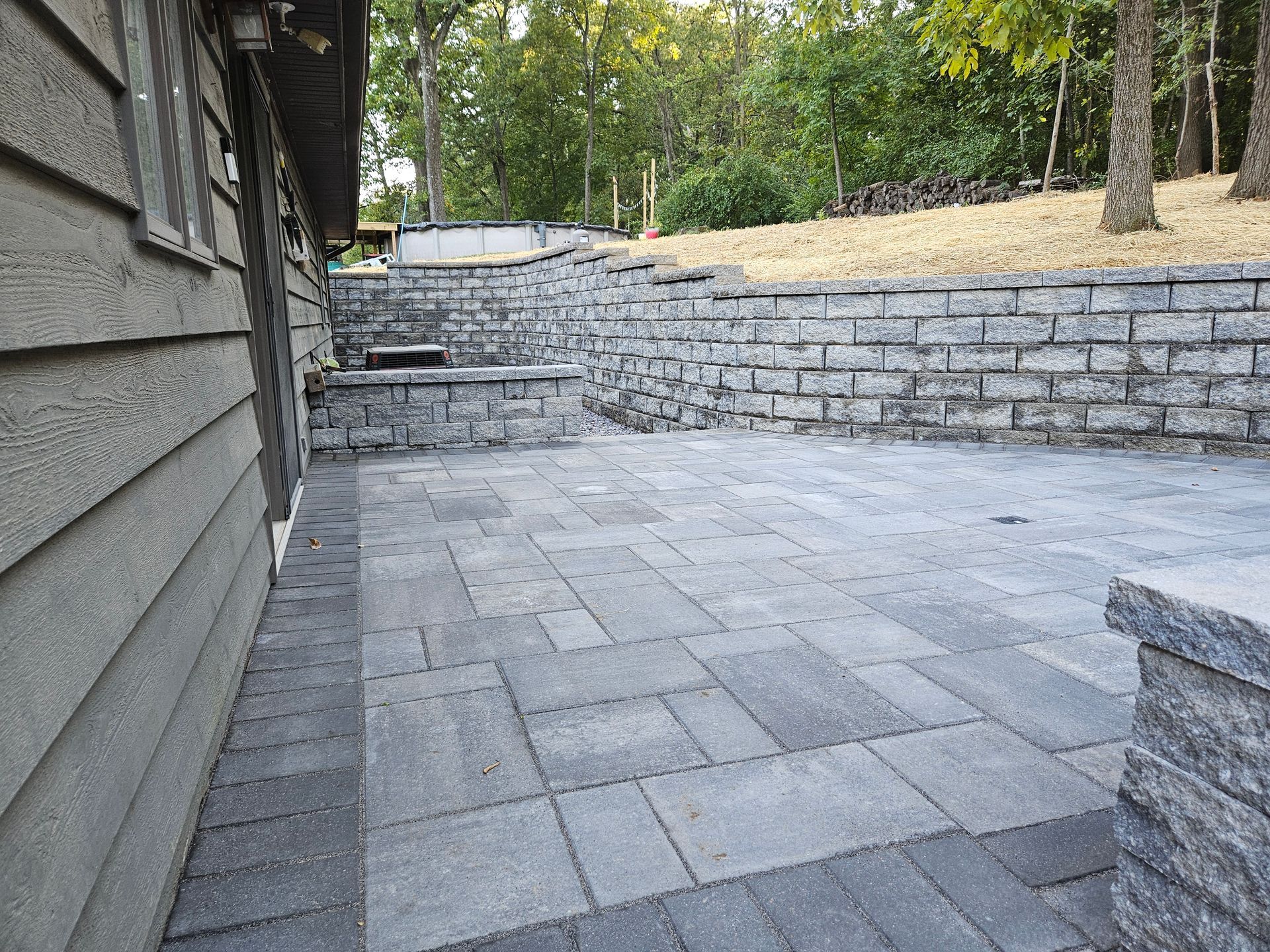 Brick patio next to a house with gray siding, leading to a retaining wall and trees.