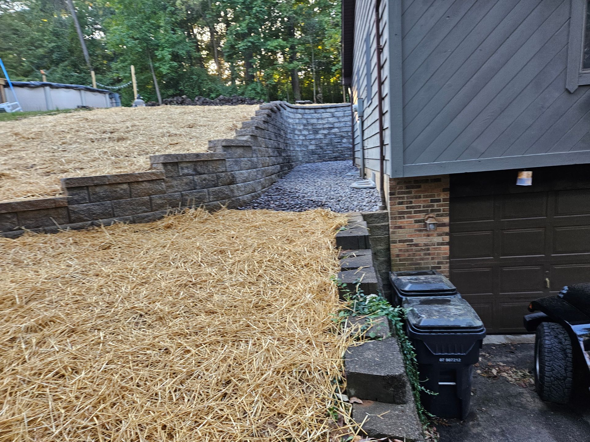 Retaining wall with steps on a sloped yard next to a house with a garage. Dry grass and a dark trash can are visible.