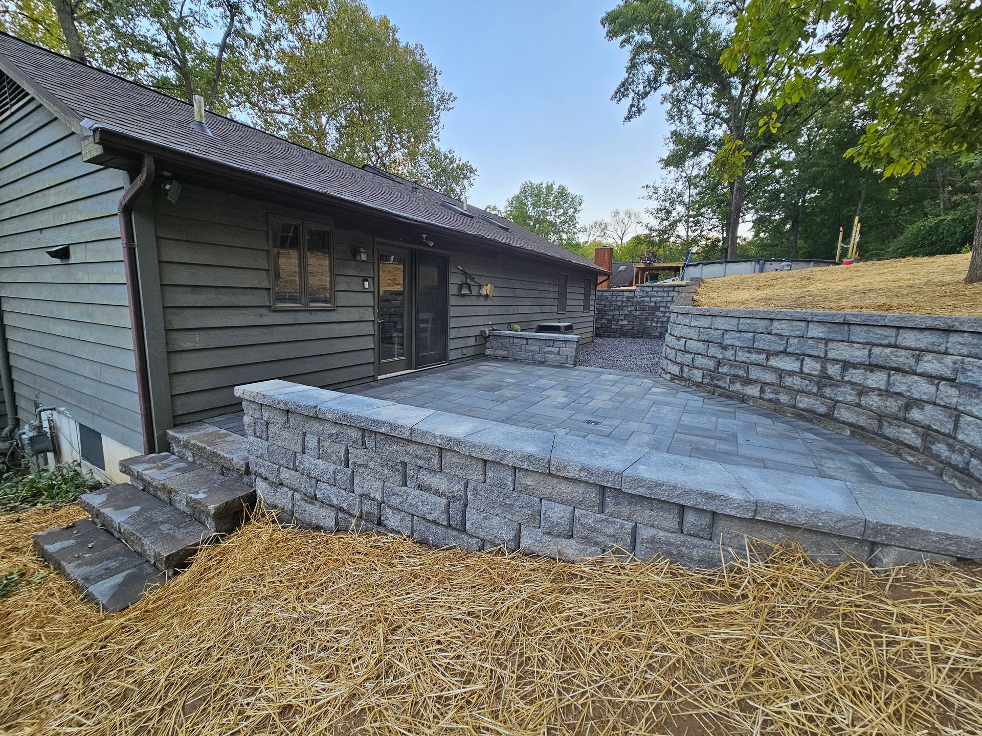 Gray house with a stone patio and retaining walls. Dry grass in the foreground, trees in the background.