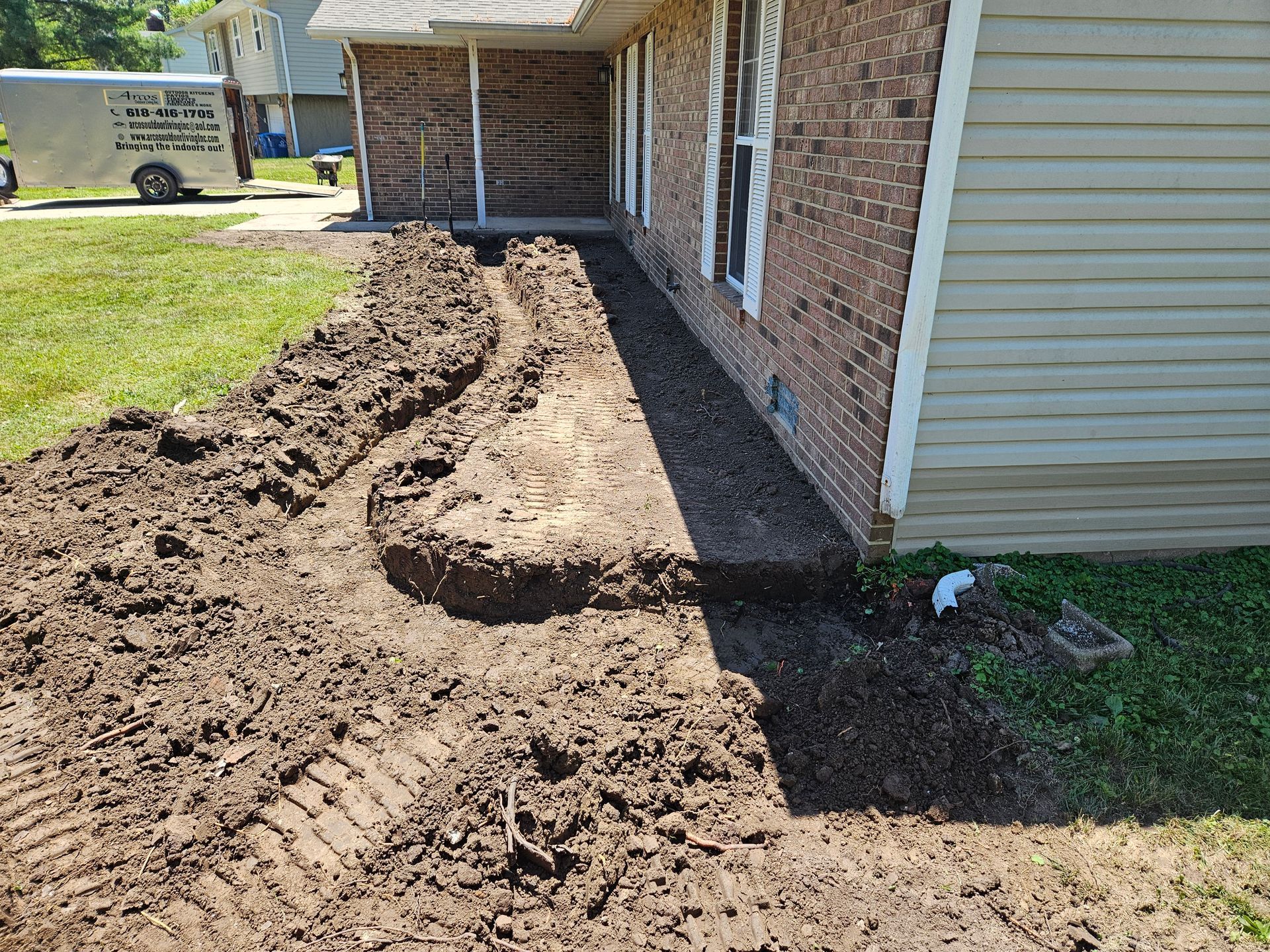 A trench dug along a brick wall of a house, with a grass lawn and a trailer in the background.