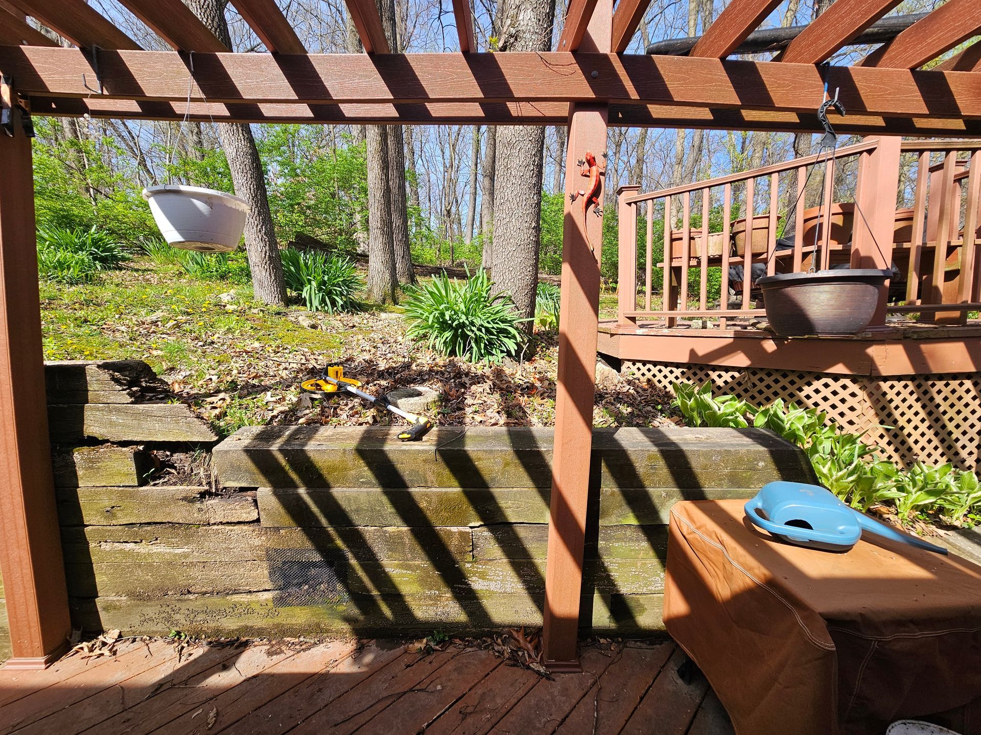 Wooden pergola casting shadows onto a retaining wall and wooden deck; potted plants and trees in the background.