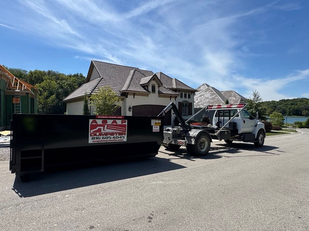 A black dumpster on a residential street being loaded by a truck, with a house and lake in the background.