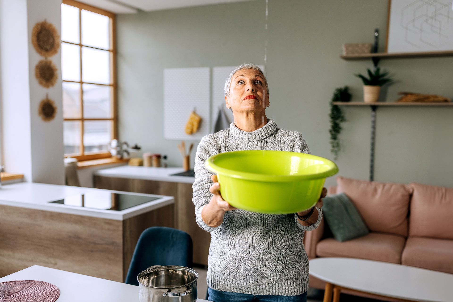 Senior woman looks at the ceiling while collecting water which leaks in the living room. Senior woman looks at the ceiling while collecting water which leaks in the living room.