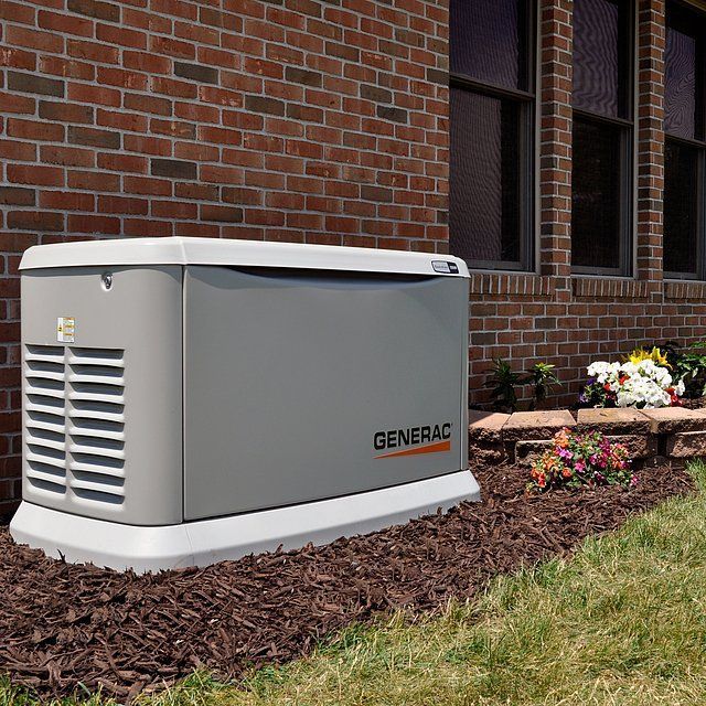 Generac generator, gray, next to a brick house with a flowerbed in front.