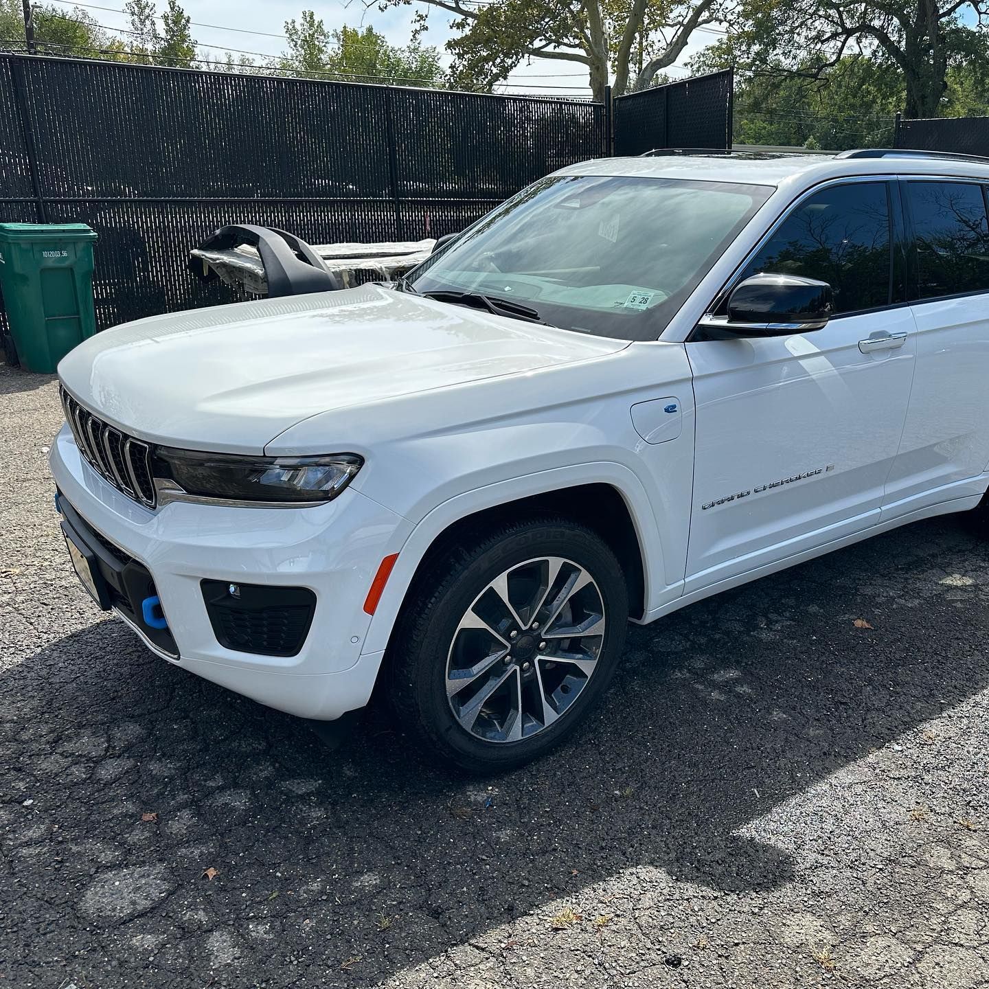 White Jeep Grand Cherokee parked outdoors, black wheels, dark tinted windows, against a fence and some equipment.