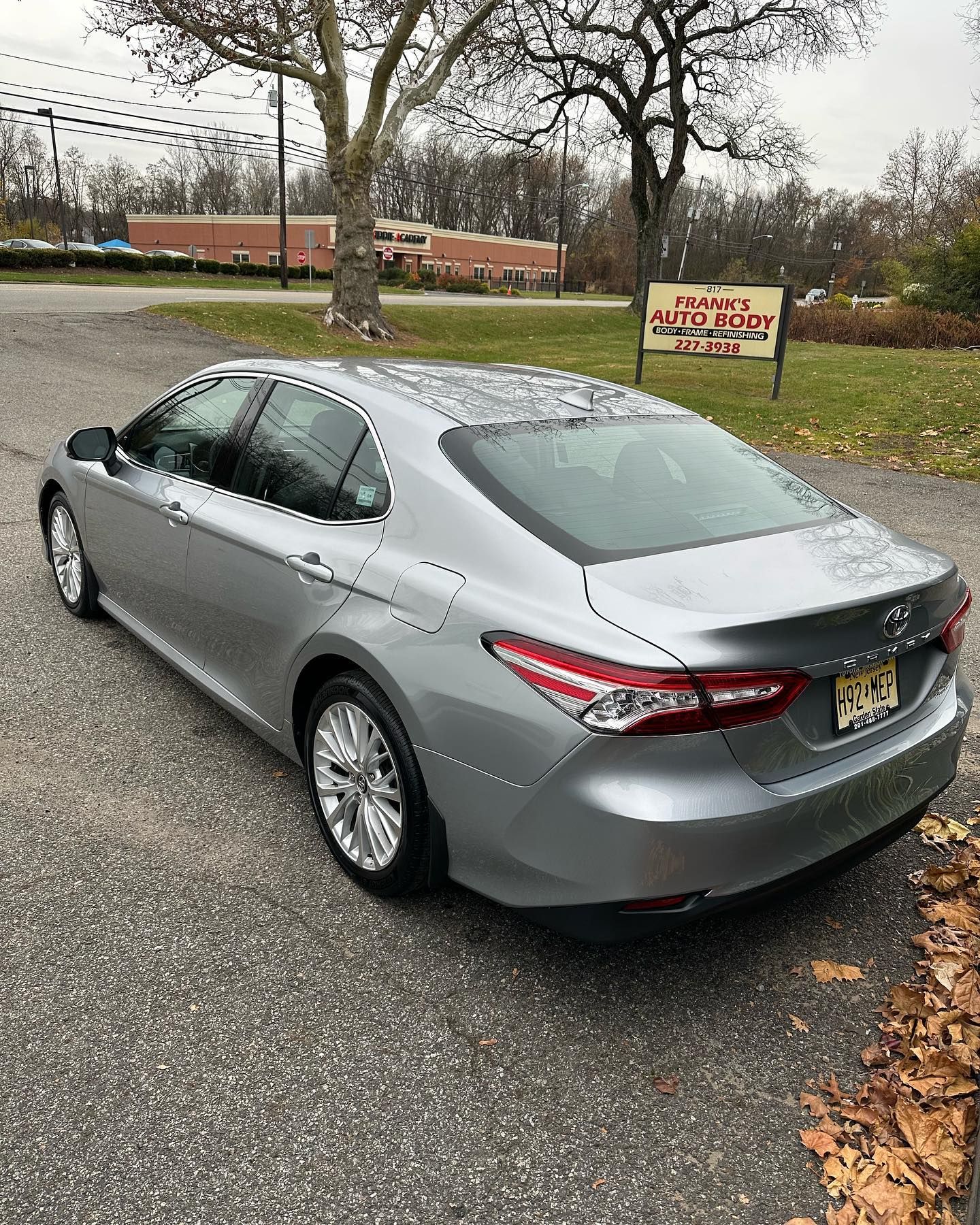 Silver Toyota Camry parked on a paved lot in front of a business with a sign. Overcast day.