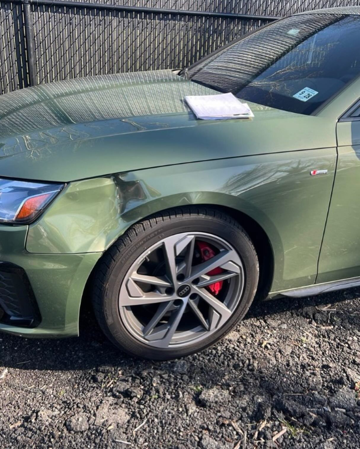 Green car with front-left fender damage, parked outdoors. A white paper sits on the hood.