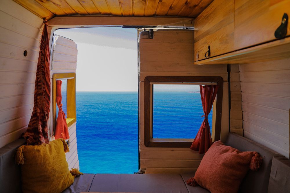 Cozy van interior with open door, overlooking blue ocean. Wooden paneling, pillows, and red curtains. — Caravan and Trailer Clinic Geelong in Breakwater, VIC