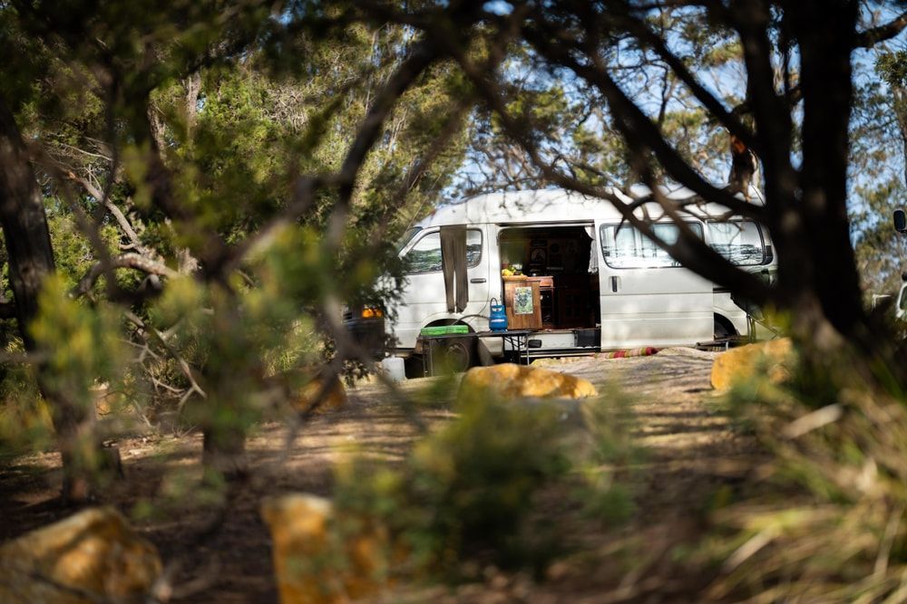 White van parked in a clearing, seen through blurred foliage. Open side door with objects visible inside. — Caravan and Trailer Clinic Geelong in Breakwater, VIC