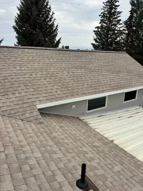 Beige shingle roof on a house, with white trim and a vent pipe. Cloudy sky and trees in the background.