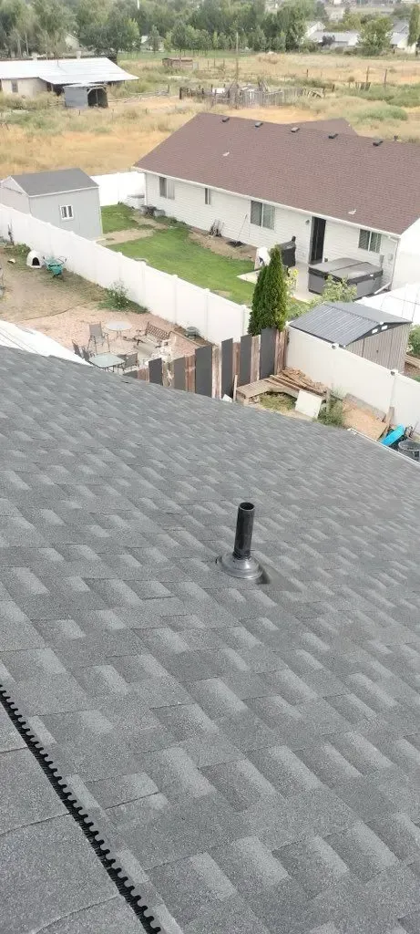 View from rooftop, gray asphalt shingles with black pipe. Background shows houses and vegetation.