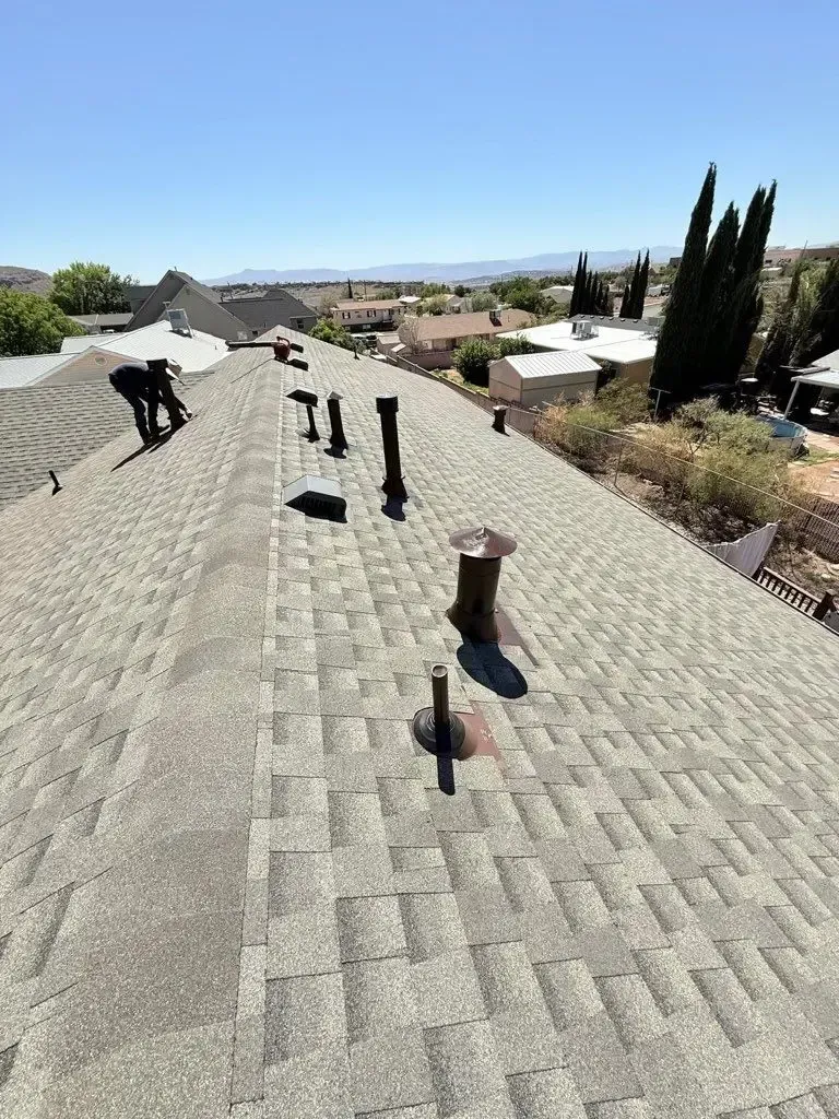 Workers on a gray shingle roof, surrounded by vents, under a blue sky, overlooking a town.